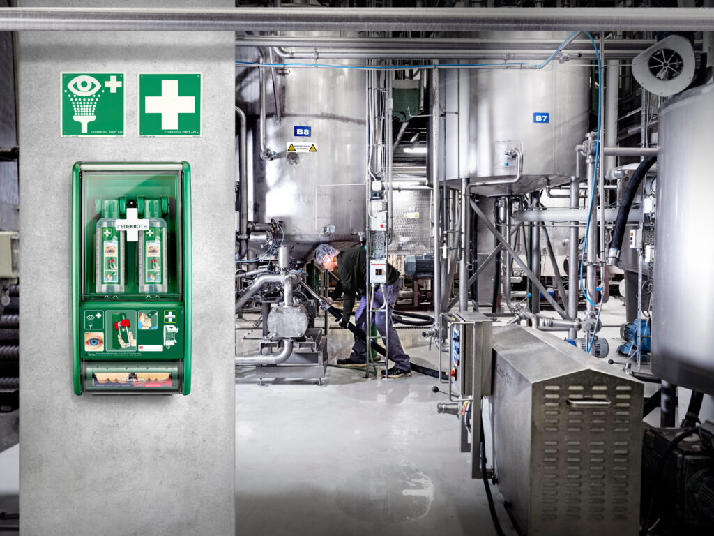 Green first aid station mounted on a wall with eye wash bottles and bandages, accompanied by green signs displaying eye wash and medical cross symbols; industrial machinery and a worker in the background.