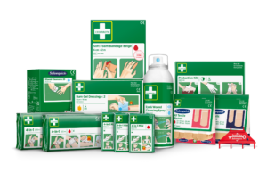 Various first aid products displayed, including wound cleansers, bandages, burn gel dressings, cleansing spray, protection kits, textile plasters, and mini kits, primarily in green packaging with illustrations and text detailing their usage.