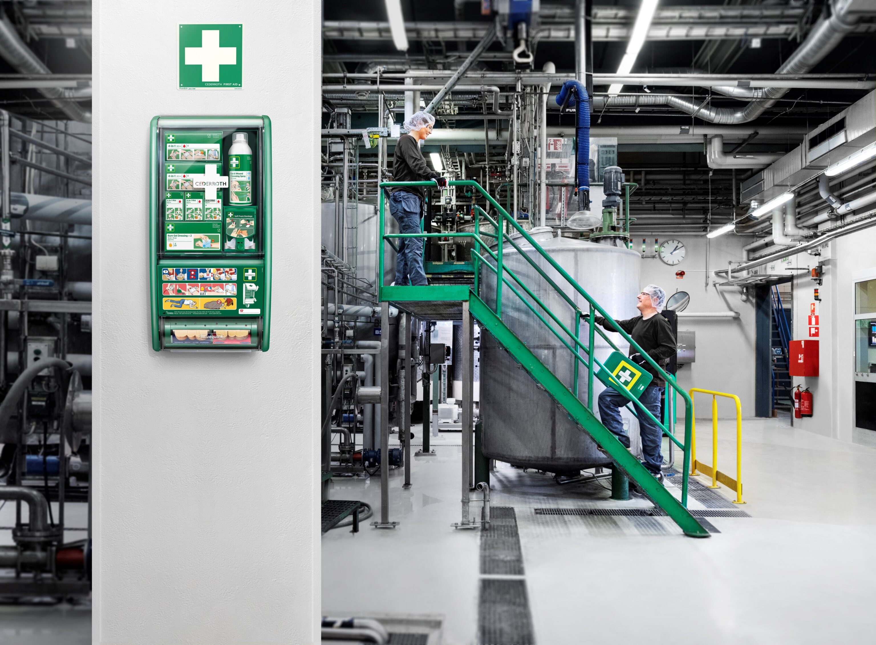 Seinällä teollisuustilassa vihreä ensiapuasema, jossa näkyy ohjeita ja ensiaputarvikkeita. Taustalla kaksi henkilöä työskentelee metallisen laitteen lähellä, toinen kantaa vihreää ensiapulaukkua portailla.