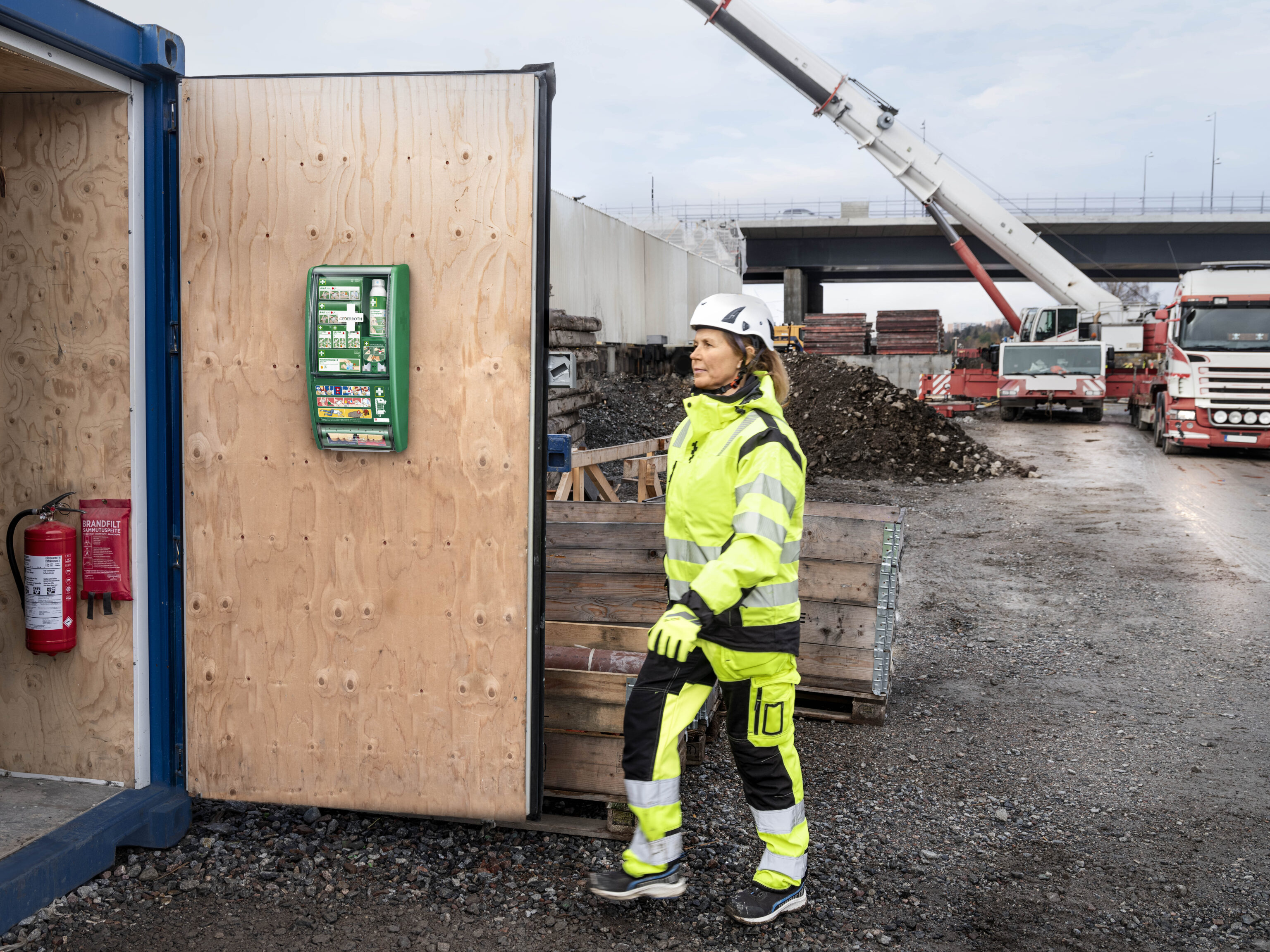 Eine Person in gelber Schutzkleidung und Helm geht an einer Baustelle entlang. Im Vordergrund ist eine offene Holztür mit einem Erste-Hilfe-Kasten und einem Feuerlöscher zu sehen, während im Hintergrund ein Kran und Lastwagen stehen.