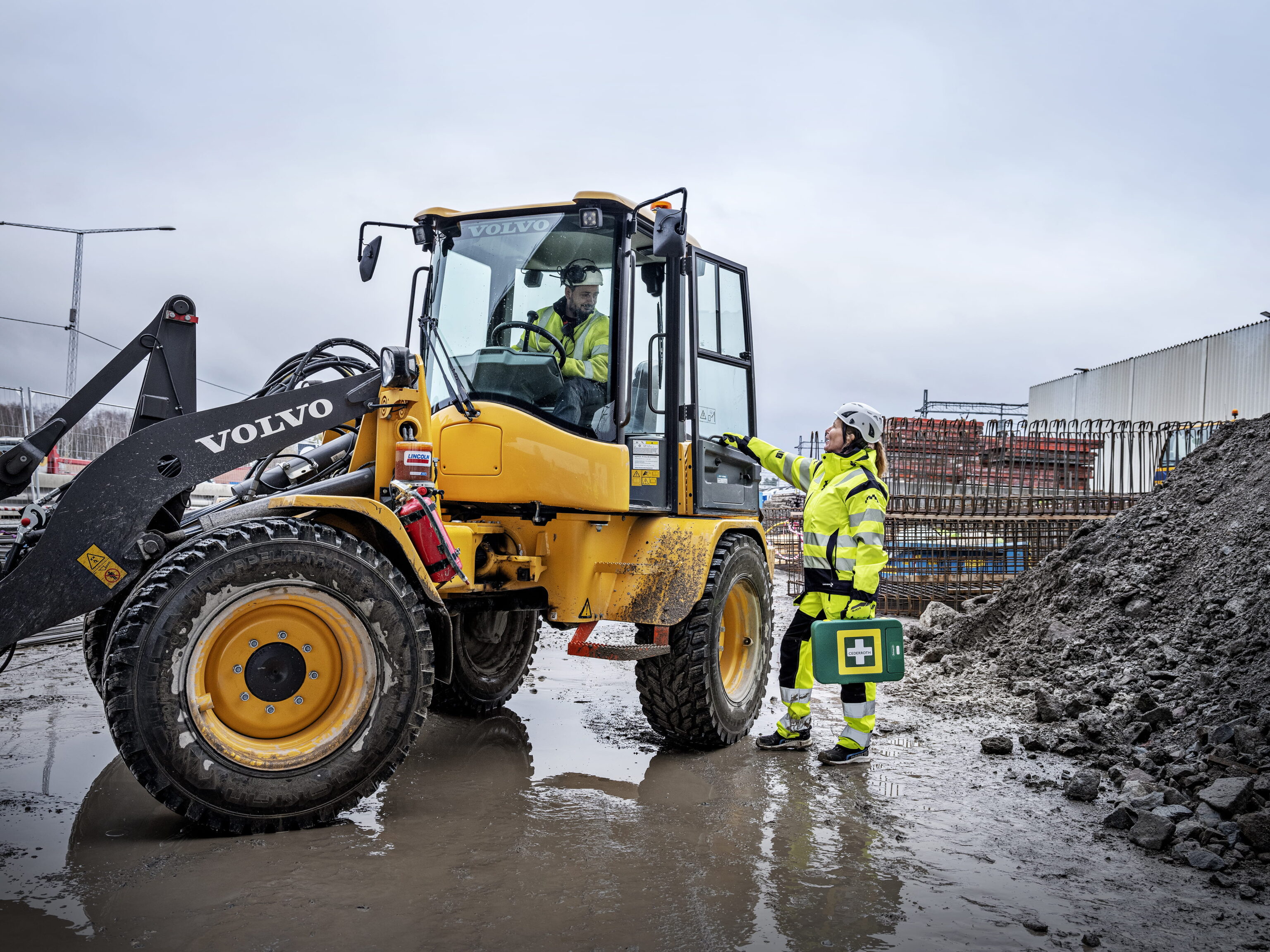 Eine Person in gelber Sicherheitskleidung und Helm steht neben einem gelben Volvo-Bagger auf einer schlammigen Baustelle und zeigt auf die Kabine, während sie eine grüne Erste-Hilfe-Box hält.