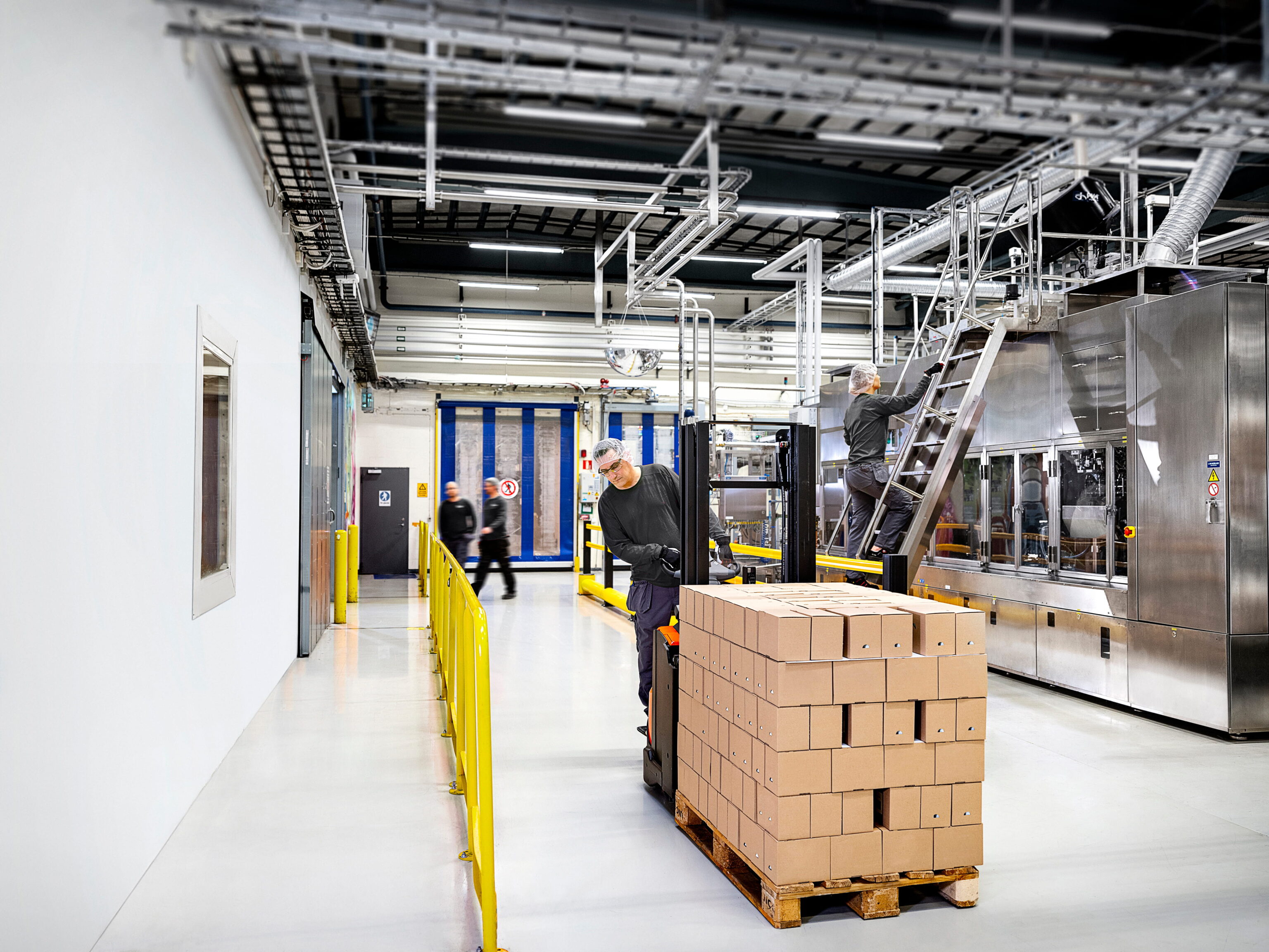 Een fabriekshal met een medewerker die een pallet met dozen verplaatst en een andere medewerker die een metalen trap beklimt naast een grote machine. In de achtergrond lopen twee mensen langs een blauwe poort.