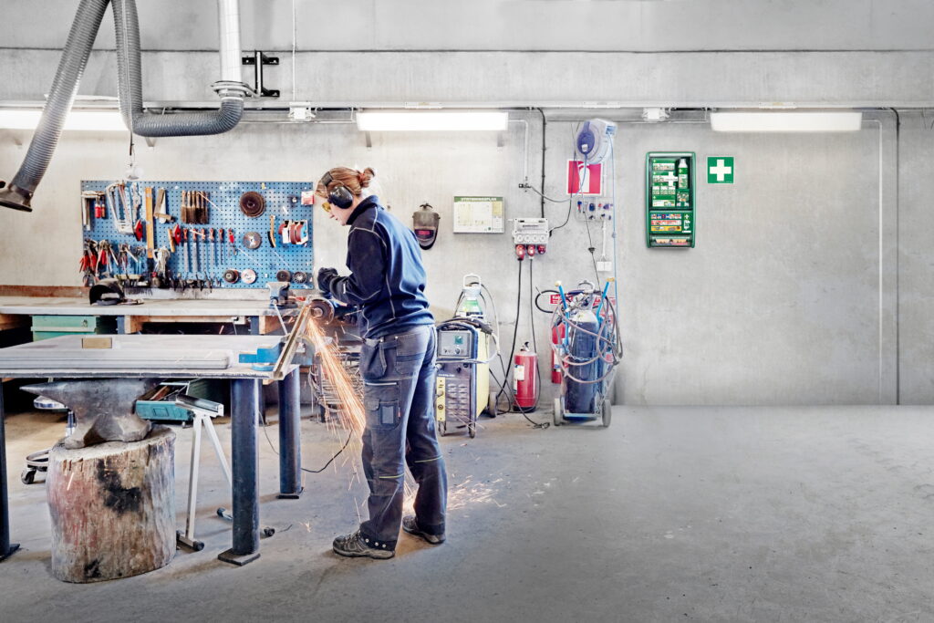 Een persoon in werkkleding slijpt met een machine met vonken in een werkplaats; gereedschap hangt aan een wandrek en veiligheidsmateriaal, zoals een EHBO-kit en brandblussers, is zichtbaar.