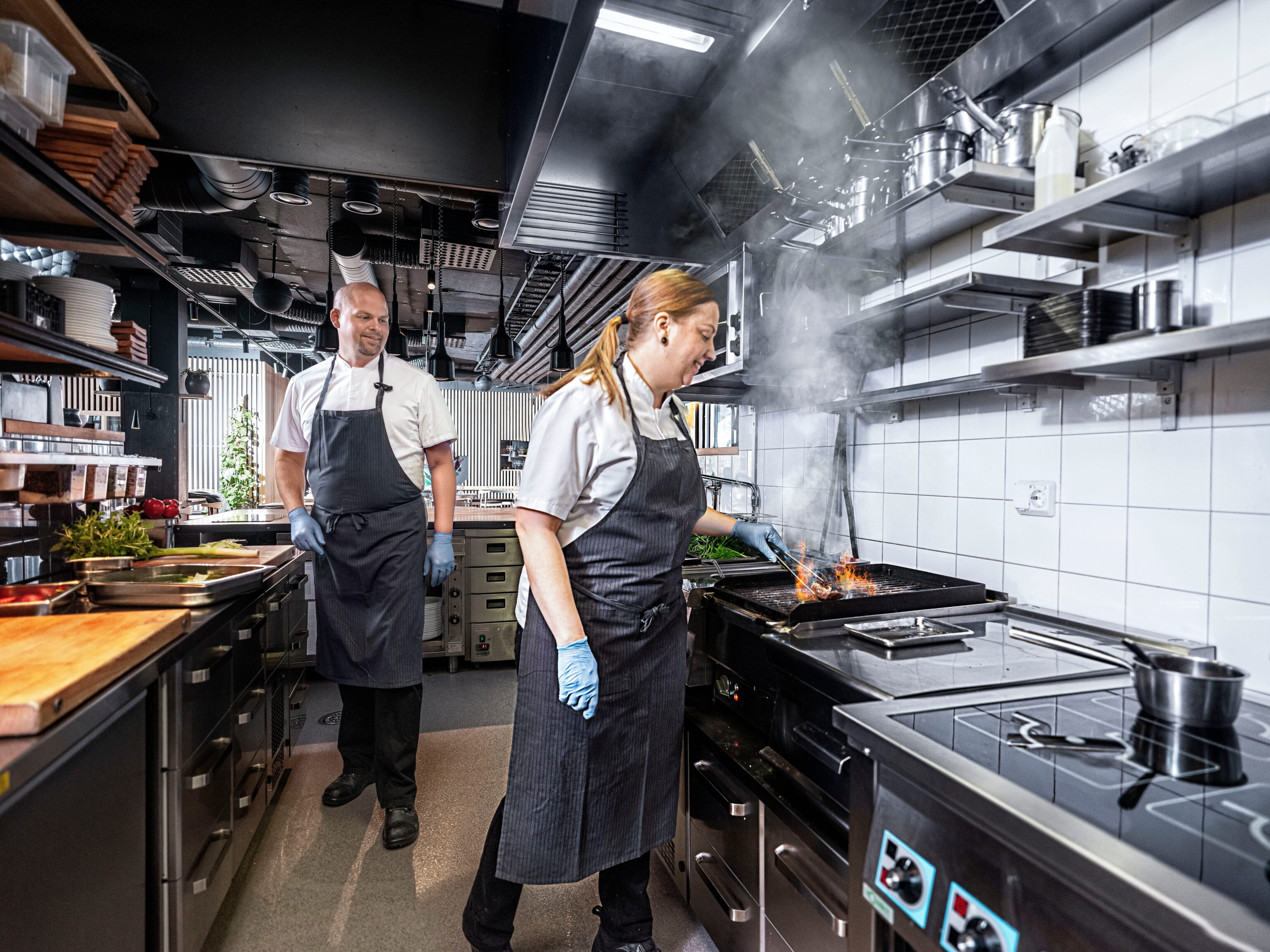 Twee koks in een professionele keuken; een vrouw bakt op een grillpan met vlammen en rook, terwijl een man op de achtergrond staat naast een werkblad met ingrediënten.