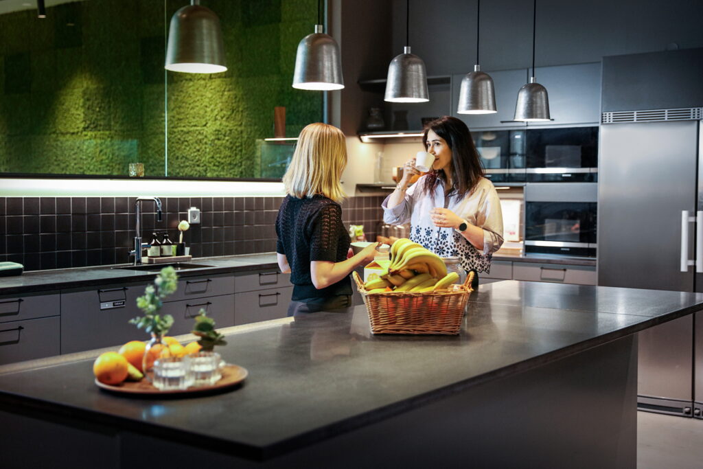 Twee vrouwen staan in een moderne keuken en drinken koffie; op het aanrecht staat een mand met bananen en een dienblad met fruit en glazen.