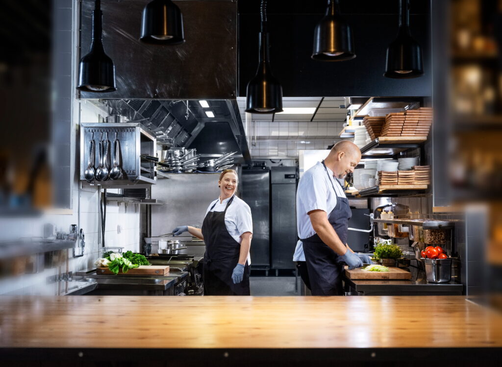 Twee koks in een professionele keuken dragen witte uniformen en zwarte schorten terwijl ze groenten bereiden; op de achtergrond hangen keukengerei en staan schappen met borden en ingrediënten.