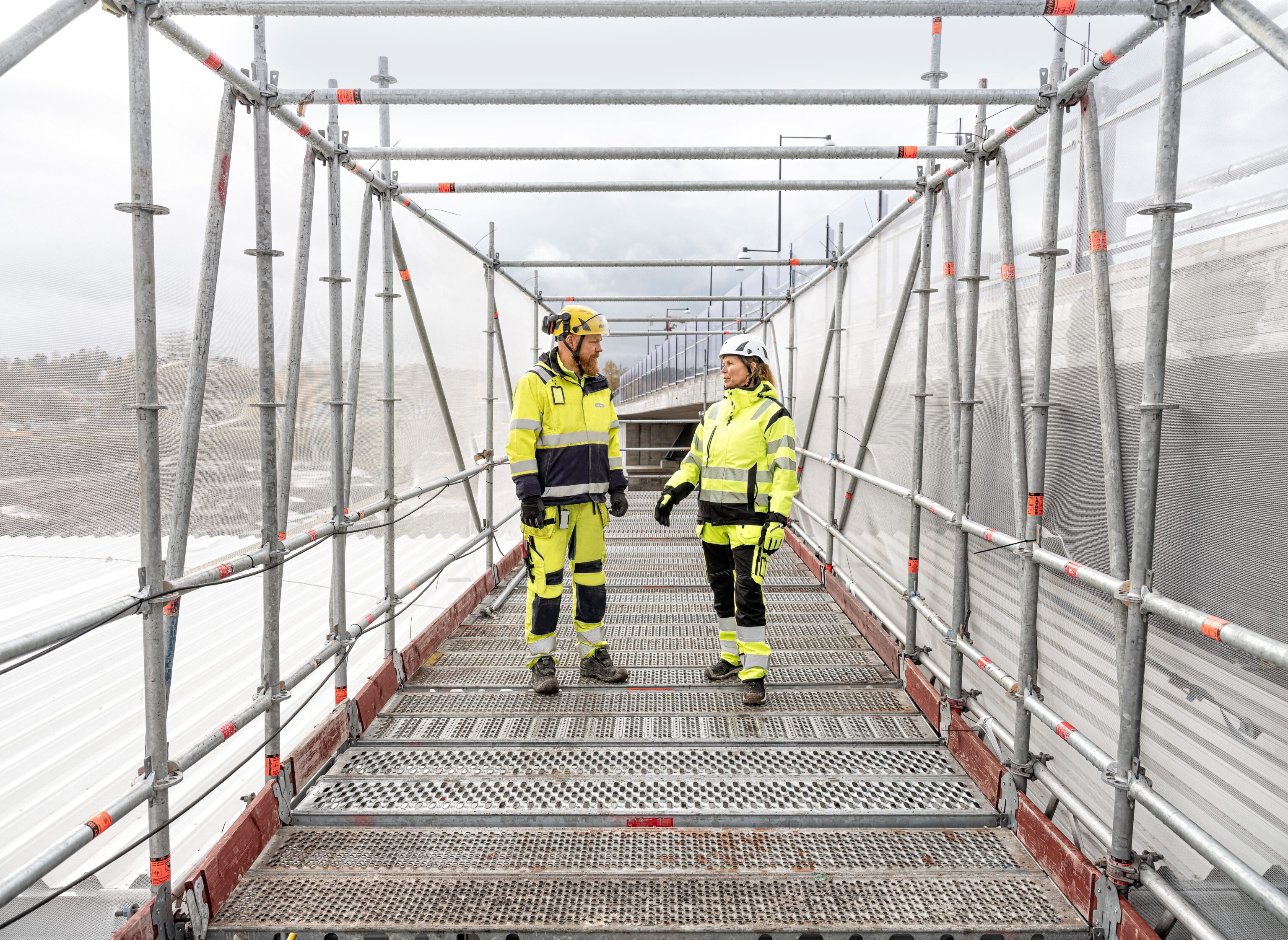 Twee bouwvakkers in felgele veiligheidskleding en helmen staan op een steiger met metalen platen, omgeven door buizen en veiligheidsnetten.