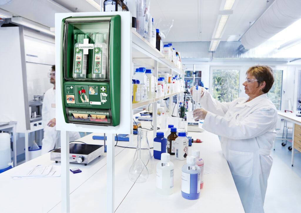 Een groene oogspoelstation van Cederroth hangt aan de muur van een laboratorium, met op de achtergrond een persoon in een witte laboratoriumjas die met reageerbuizen werkt te midden van flessen met chemicaliën.