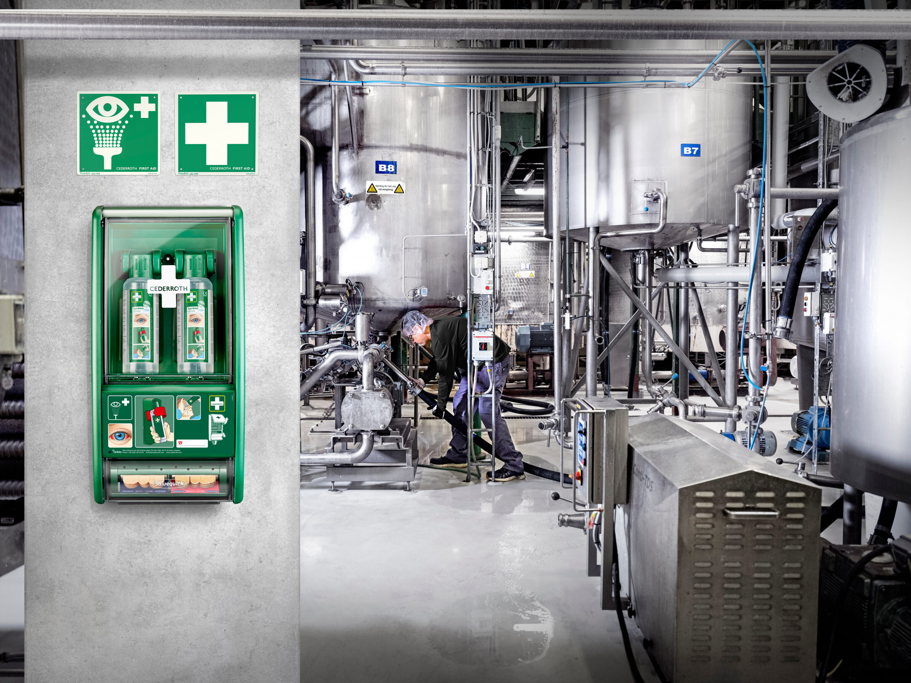 Na ścianie w industrialnym zakładzie znajduje się zielony zestaw pierwszej pomocy marki Cederroth z oznaczeniami bezpieczeństwa, obok dwóch zielonych tabliczek z symbolem oka i krzyża. W tle pracownik obsługuje maszynę wśród metalowych zbiorników i rur.