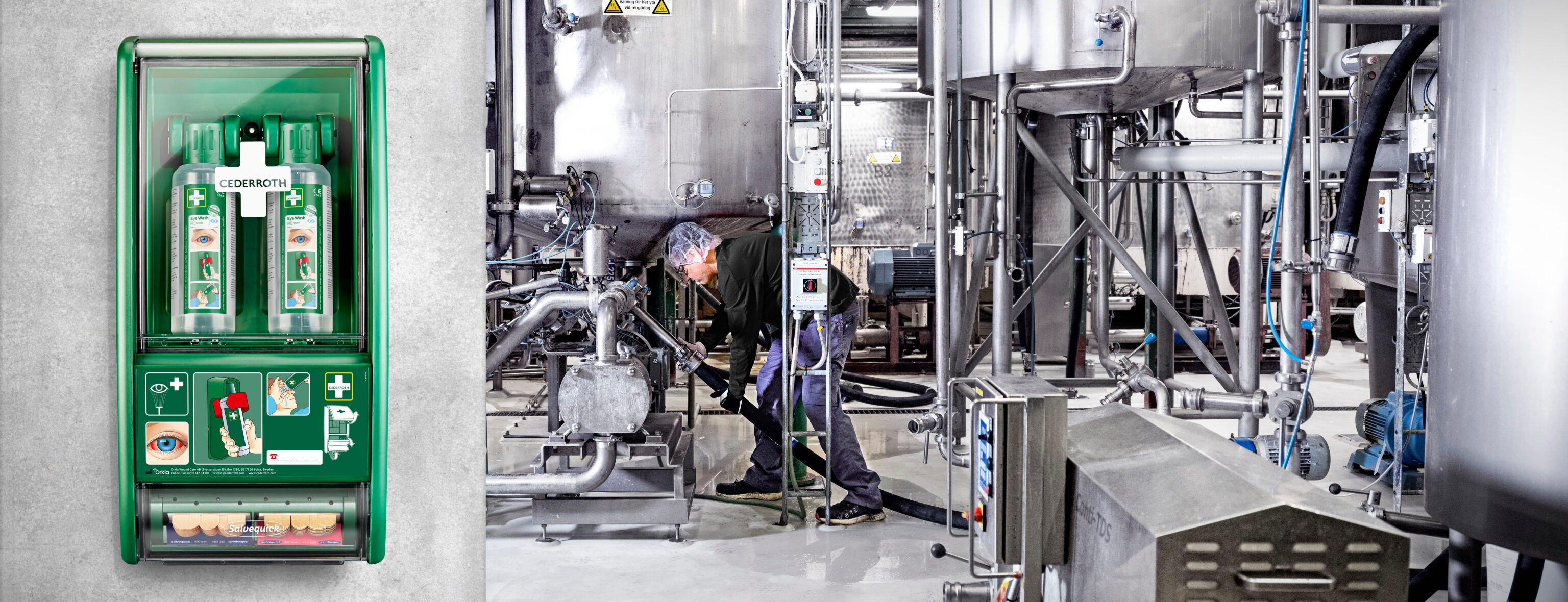 Första hjälpen-station med ögondusch och plåsterdisplay monterad på en grå vägg. I bakgrunden syns en industriell miljö med metalltankar och rörsystem, där en person arbetar med utrustning.